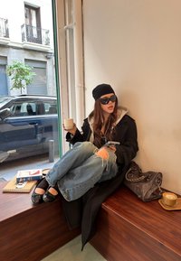 Svart overcoat med grå hettegenser, slitte blå jeans, svarte flate sko, solbriller og en beanie. Holder en kaffekopp, med telefon ved siden av.