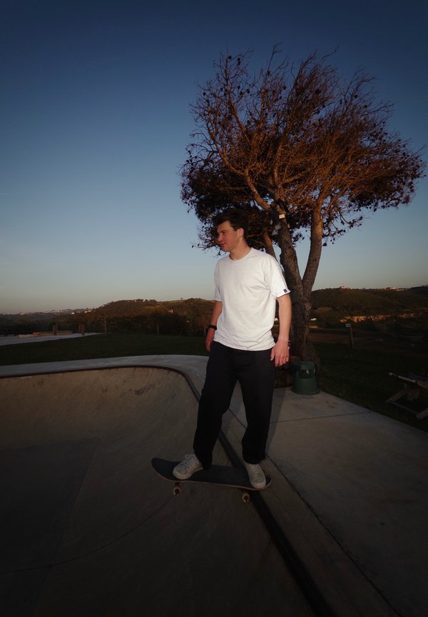 Ein junger Mann im weißen Hemd fährt mit dem Skateboard eine geschwungene Betonrampe in einem Skatepark im Freien bei Sonnenuntergang hinunter.