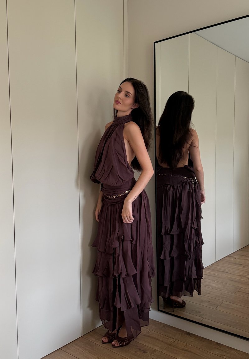 Mujer con vestido sin mangas, en capas y de color púrpura oscuro, de pie cerca de un espejo que refleja su espalda, en una habitación con paredes claras y suelo de madera.