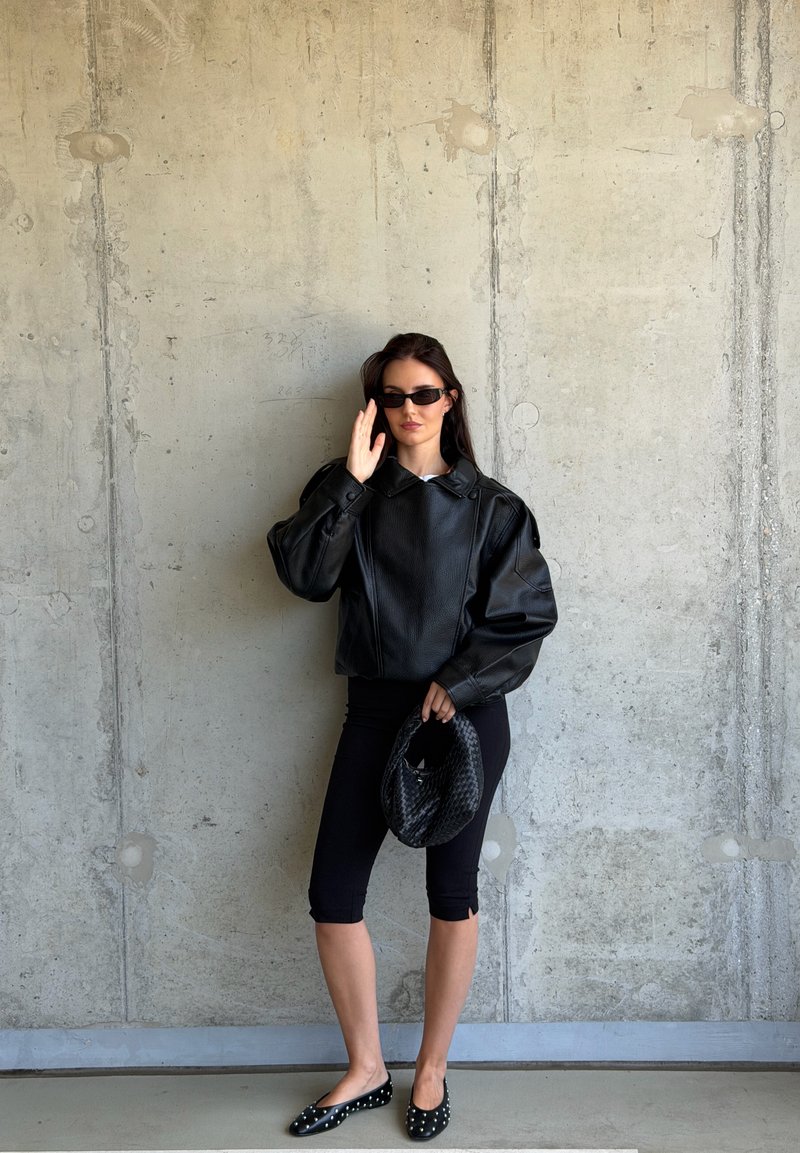 Mujer con chaqueta de cuero negra, leggings negros recortados, zapatos planos con tachuelas y gafas de sol, sosteniendo un bolso negro tejido contra una pared de hormigón.