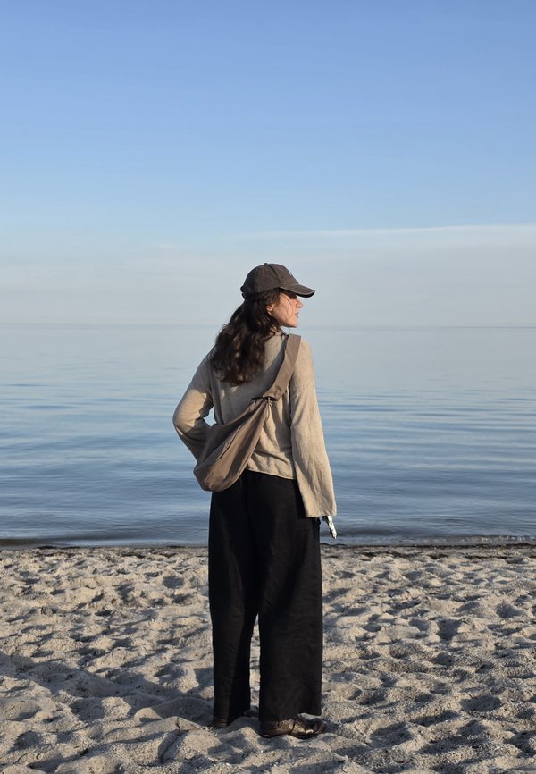 Donna con cappellino e top beige in piedi su una spiaggia sabbiosa che guarda il mare calmo sotto un cielo azzurro terso.