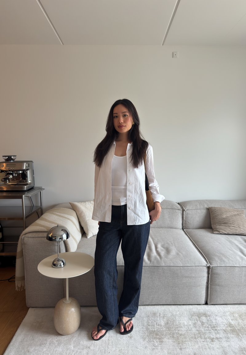 Une femme se tient dans un salon moderne, vêtue d'une chemise blanche, d'un top blanc, d'un jean foncé et de sandales, à côté d'un canapé, d'une lampe et d'une table basse.