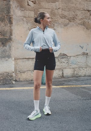 Mujer joven con ropa deportiva de pie en la calle, subiendo una sudadera azul clara con cremallera, mirando hacia la derecha, con un fondo de pared de piedra texturizada.