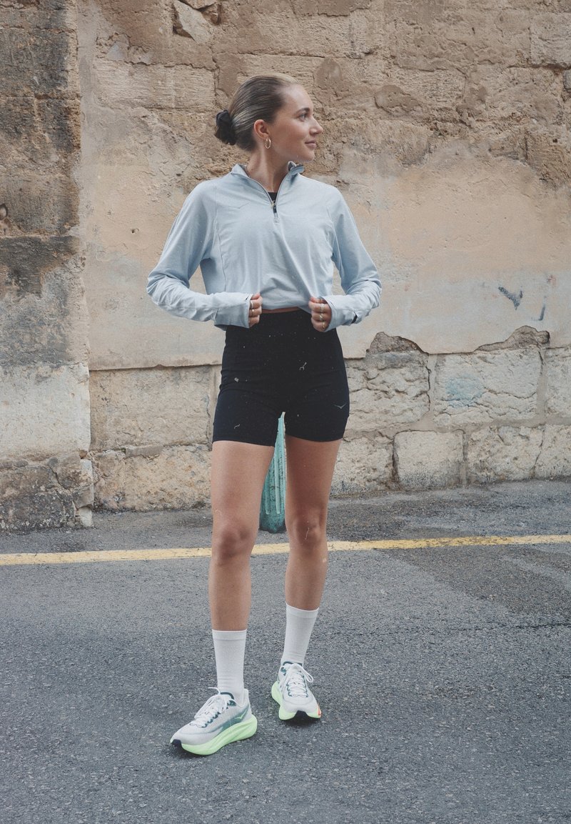 Jeune femme en tenue de sport debout dans la rue, remontant un haut zippé bleu clair, tournée vers la droite, devant un mur en pierre texturé.