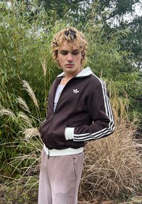 Veste marron à fermeture éclair avec des rayures blanches sur les manches, col blanc, associée à un pantalon de survêtement rose clair, sur fond d'herbe verte et sèche.