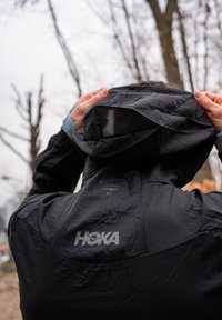 Casaco leve preto com capuz, apresentando uma superfície texturizada e o logótipo HOKA nas costas. O design é minimalista e funcional.