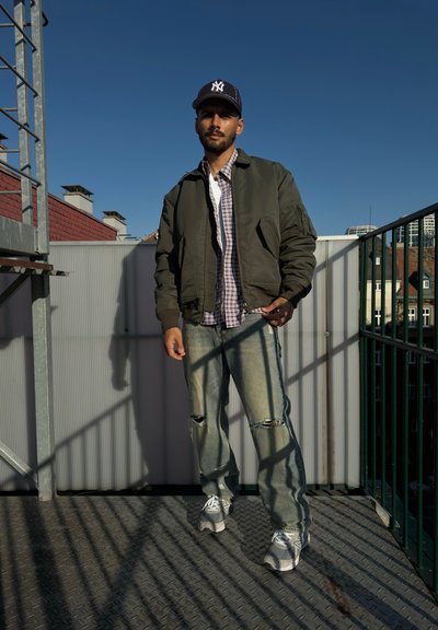 Chaqueta bomber verde sobre una camisa a cuadros, jeans claros desgastados y zapatillas grises. Gorra negra con logo blanco, de pie sobre una plataforma de metal.