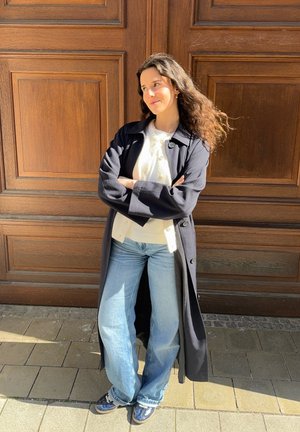 Abrigo negro largo sobre una camisa blanca, combinado con unos vaqueros de pierna ancha de color azul claro y zapatillas navy, de pie contra un fondo de madera.