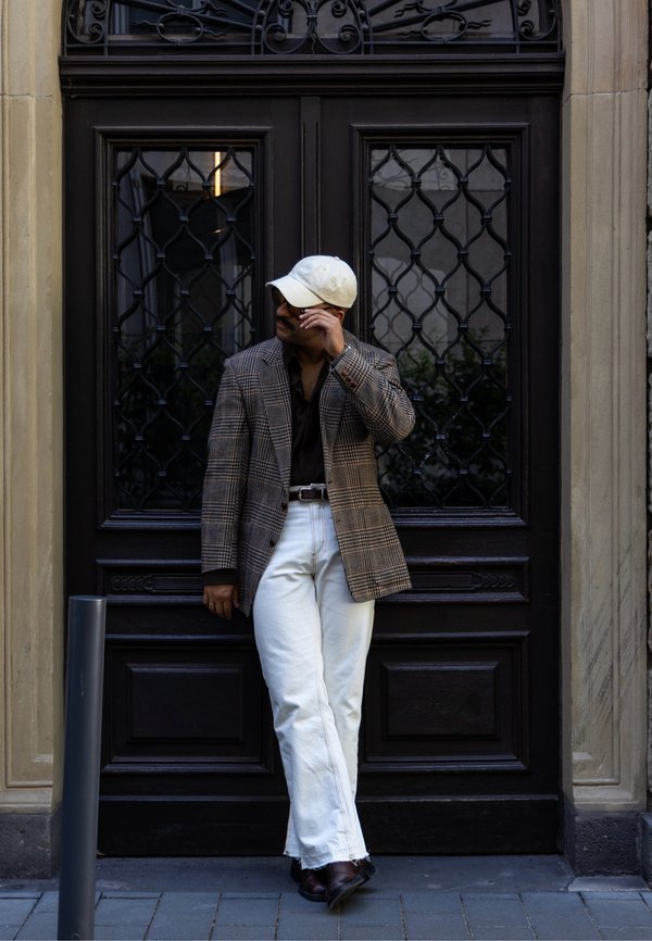 Hombre con gorra blanca, blazer marrón a cuadros, camisa negra y pantalones blancos apoyado en una puerta negra ornamentada, ajustándose las gafas de sol.