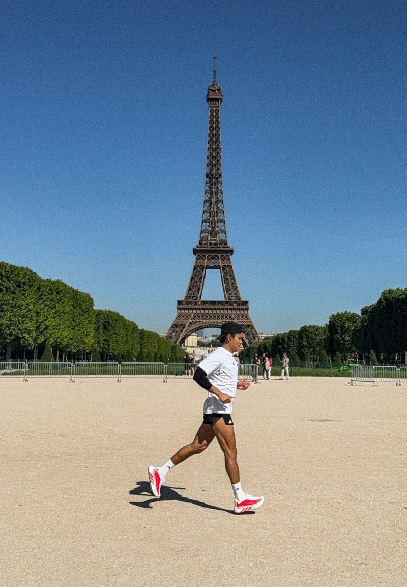 Homme courant sur un chemin sablonneux avec la tour Eiffel en arrière-plan sous un ciel bleu clair dans une zone de parc avec des arbres et des barrières.