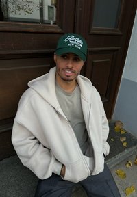 Ανοιχτό μπεζ φούτερ με φερμουάρ πάνω από γκρι πουκάμισο, σκούρες ριγέ παντελόνες και πράσινο καπέλο μπέιζμπολ με λευκό κείμενο, καθισμένος σε πέτρινα σκαλιά.