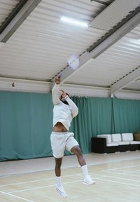 Joueur en tenue de sport gris clair frappe une balle de tennis avec une raquette violette. Terrain intérieur avec des rideaux verts et des sièges blancs en arrière-plan.