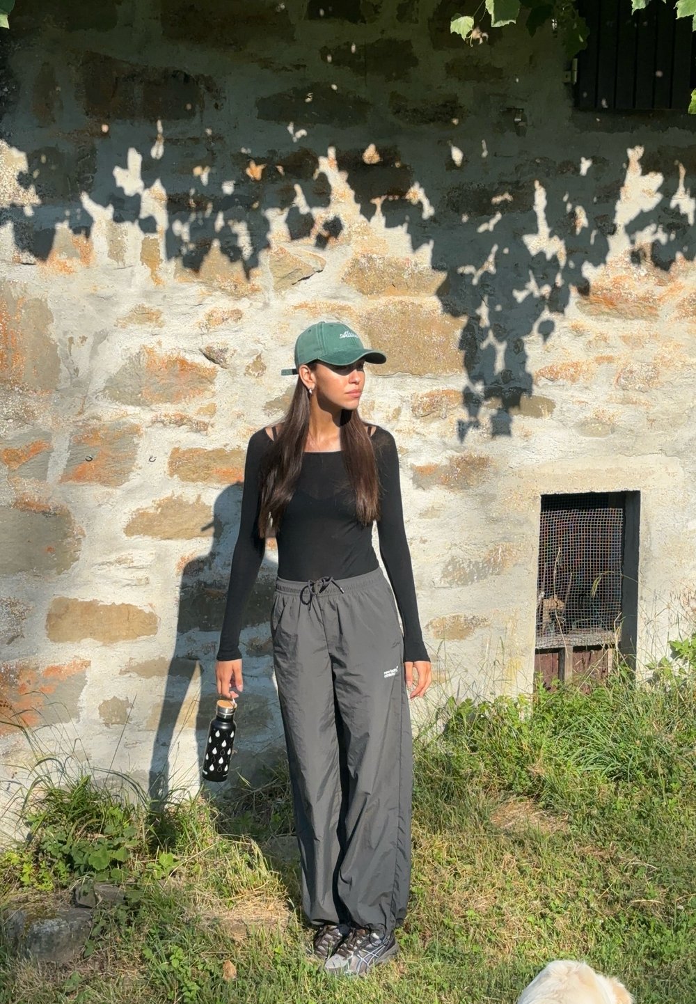 Top negro de manga larga, pantalones grises holgados, gorra verde, sosteniendo una botella de agua negra con lunares blancos, de pie frente a una pared de piedra.