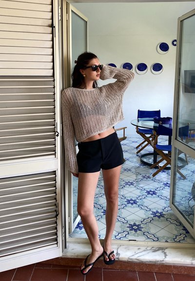 Suéter de punto beige claro, pantalones cortos negros y chanclas. El ambiente presenta azulejos estampados y puertas correderas abiertas.