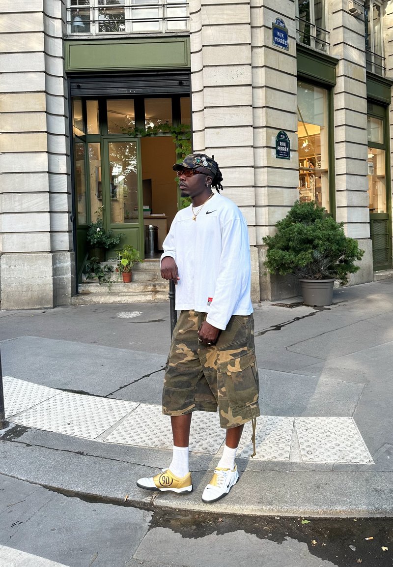 Långärmad vit skjorta med svart logotyp, kamouflage cargo-shorts, gula och vita sneakers. Tillbehör inkluderar solglasögon och en mönstrad keps.