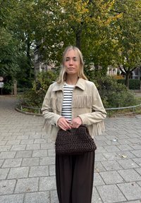 Giacca beige in suede con dettagli a frange, camicia a righe nera e bianche, borsa intrecciata marrone e pantaloni scuri in un ambiente all'aperto.