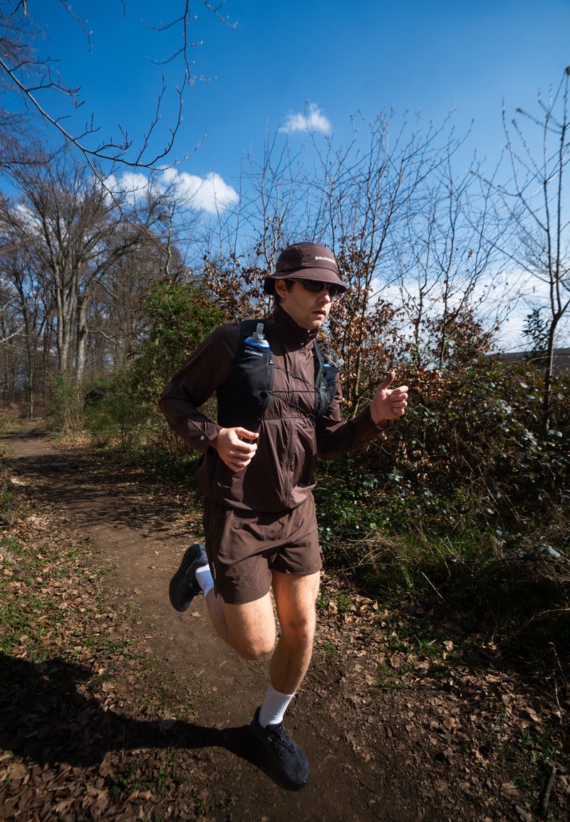 Muž oblečený do hnědého sportovního oblečení a čepice běží po lesní stezce pod jasně modrou oblohou s holými stromy a suchým listím.