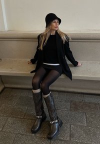 Vysoké hnědé kožené boty se stříbrnými doplňky a pletenými manžetami, nošené s černými punčocháči a černým svetrem. Osoba sedí na kamenné lavici.