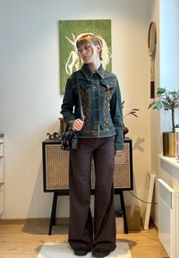 Femme debout à l'intérieur portant une veste en jean et un pantalon marron à fines rayures, avec un sac à main noir, près d'une peinture verte et de plantes d'intérieur.