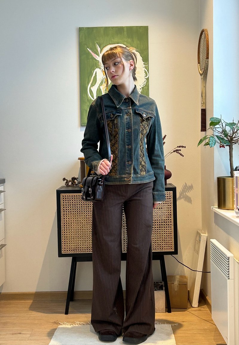 Femme debout à l'intérieur portant une veste en jean et un pantalon marron à fines rayures, avec un sac à main noir, près d'une peinture verte et de plantes d'intérieur.