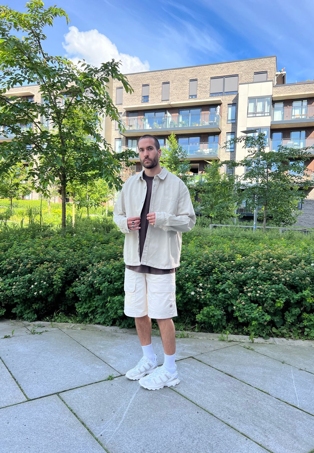 Giacca oversized beige chiaro sopra una maglietta scura, pantaloni cargo bianchi, sneakers bianche, in una posizione casual in un ambiente verde all'aperto.