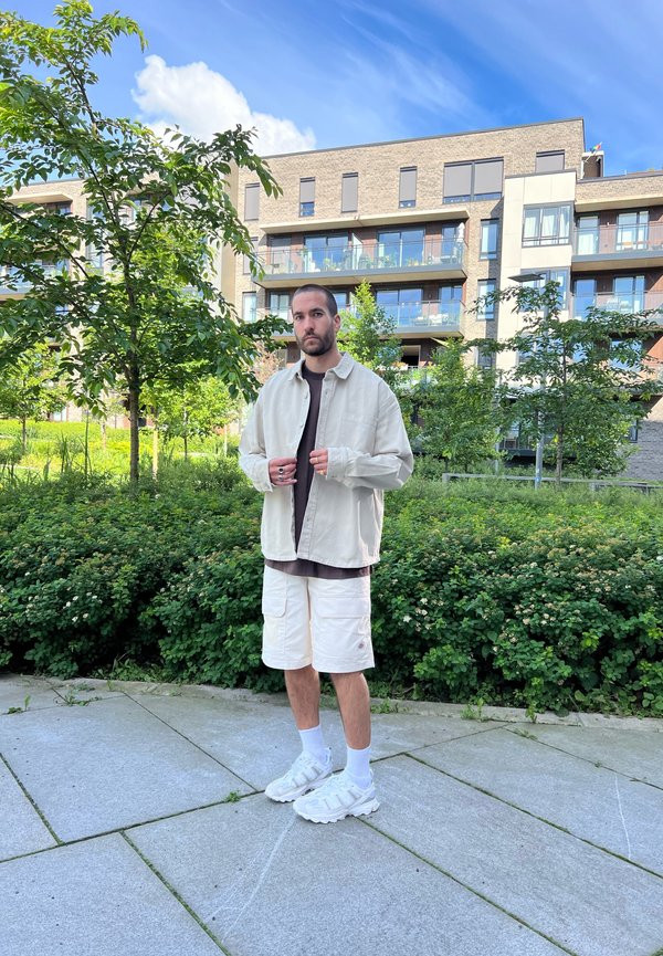 Giacca oversized beige chiaro sopra una maglietta scura, pantaloni cargo bianchi, sneakers bianche, in una posizione casual in un ambiente verde all'aperto.