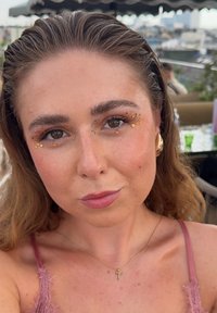 Kvinne med bølgete brunt hår, iført en rosa kjole med blonder. Subtil sminke fremhever; gylden aksenter rundt øynene og i øredobber.