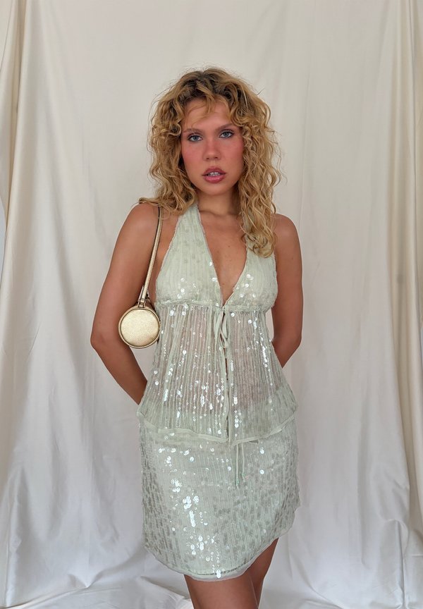 Young woman with curly blonde hair wears a light green sequined dress and holds a small round gold purse on her shoulder.