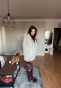 Femme portant un manteau en fausse fourrure blanc, des bottes bordeaux et un sac à main, debout à l'intérieur près d'une table avec un chat roux et des jeux de société.