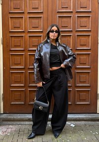 Chaqueta de cuero marrón brillante sobre una blusa negra, combinada con pantalones negros sueltos y tacones negros puntiagudos. Bolso negro con detalles dorados.