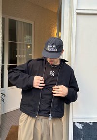 Sort lynlåstrøje over en sort T-shirt med hvidt logo, og beige bukser. Tilbehør inkluderer ringe og en sort kasket med logo.