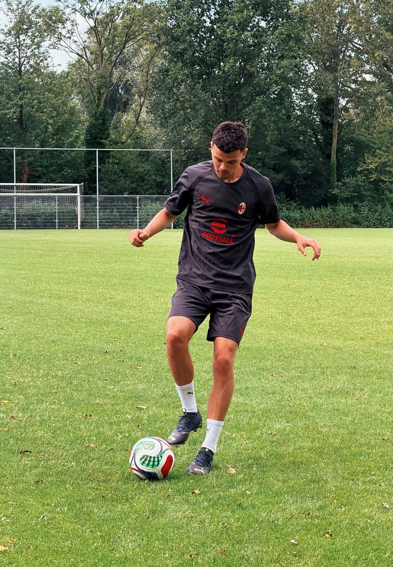 Jeune homme portant une tenue de sport noire contrôle un ballon de football sur un terrain en herbe avec un but et des arbres en arrière-plan.