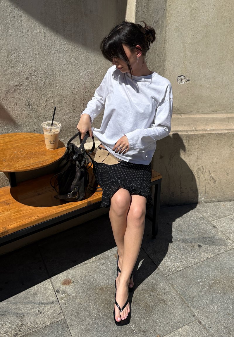Femme en chemise blanche à manches longues et jupe noire assise sur un banc, fouillant dans un sac noir, avec un café glacé sur une table en bois.