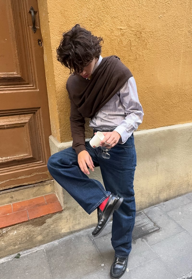 Jong persoon die op één been staat tegen een gele muur, een witte beker en een bril vasthoudend, gekleed in een blauwe spijkerbroek, een bruine sjaal en zwarte schoenen met rode sokken.