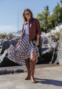 Καφέ δερμάτινες μπότες ως το γόνατο, μπορντώ κοντομάνικο μπουφάν και μία ρυθμισμένη φούστα καρό με ένα λευκό μπλουζάκι με γραφικά, σε θέση πάνω σε βραχώδη επιφάνεια.