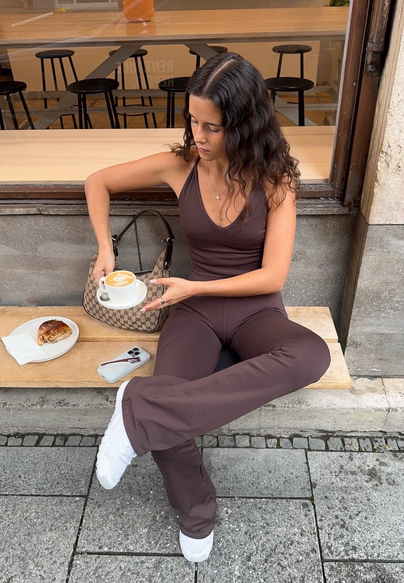 Tight-fitting, marrone tuta atletica con scollatura a V. Sneakers bianche, in mano un latte. Una borsa Gucci e una pasticceria danese su un piatto vicino.