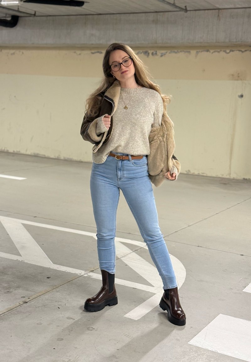 Brown shearling jacket over a light grey sweater, light blue jeans, and dark brown platform ankle boots in a parking garage setting.