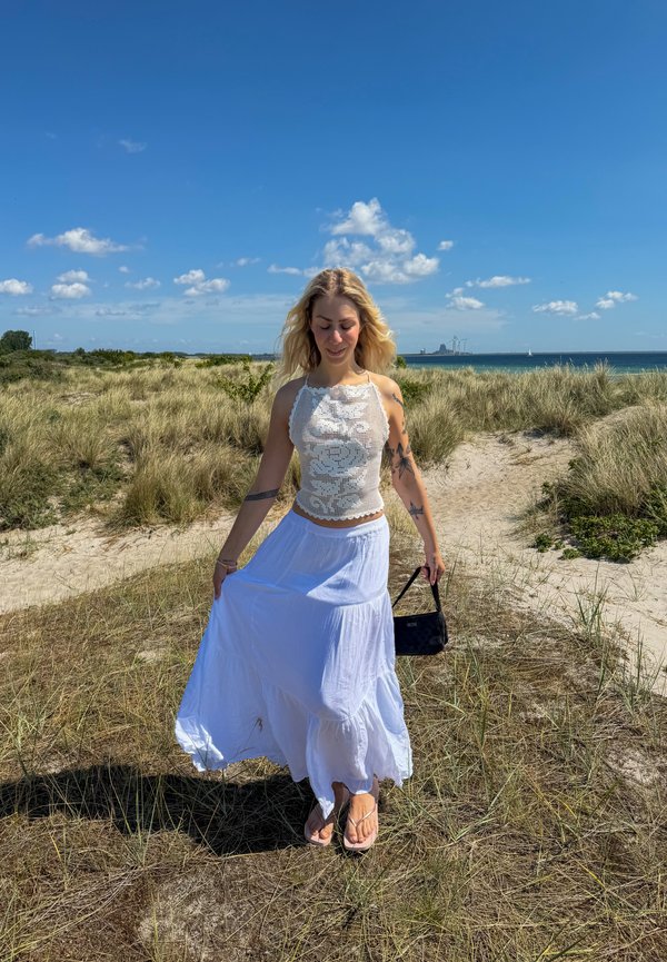 Vrouw met blond haar in een wit gehaakt topje en lange witte rok die loopt op een zanderig pad door hoog gras aan zee onder een heldere blauwe lucht.
