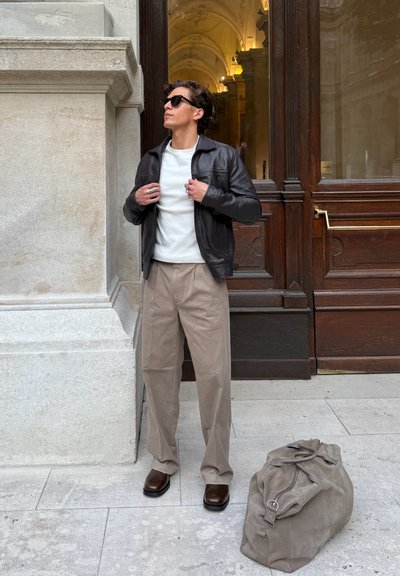 Chaqueta de cuero negra, camisa blanca, pantalones beige de pierna ancha, zapatos marrones. Bolsa gris suave en el suelo, situada cerca de una puerta de madera ornamentada.