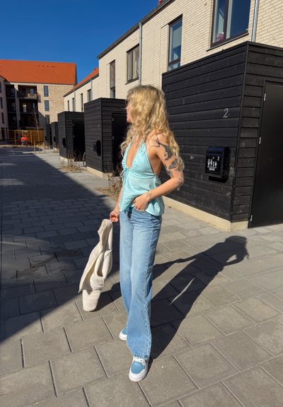 Mujer con pelo largo y rubio y tatuajes que lleva una camiseta sin mangas azul claro, vaqueros y zapatillas, de pie en un camino pavimentado junto a edificios modernos.