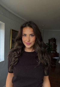 Joven con cabello largo y ondulado de color castaño, vistiendo una camiseta oscura de manga corta, de pie en el interior cerca de una ventana y muebles de madera.