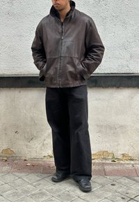 Brown leather jacket with a high collar, front zip, and two side pockets, paired with loose black trousers and black shoes.