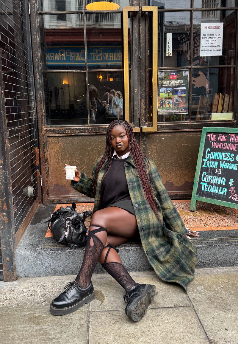 Oversized coat in plaid green and yellow, black sweater, and skirt. She wears patterned tights and black platform shoes, holding a cup.