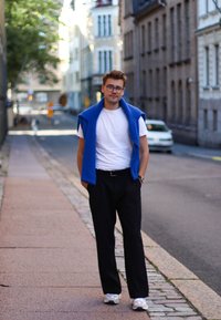 Kényelmes öltözet: fehér póló, kék pulóver a vállon, fekete nadrág és szürke sportcipő. Városi utcai környezet.