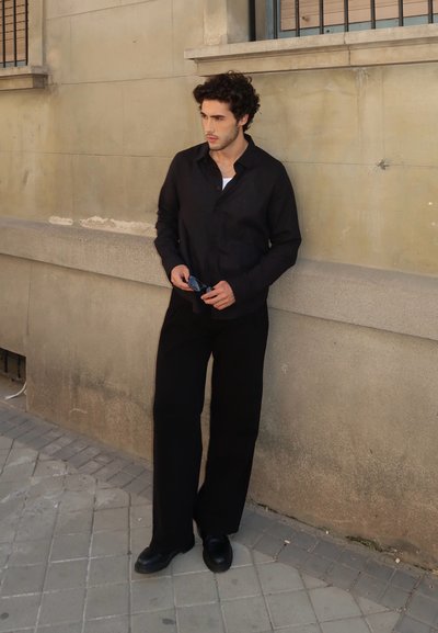 Camisa negra de botones y pantalones anchos negros, ambos hechos de tela lisa. Zapatos negros y gafas de sol en la mano. Fondo de pared neutro.