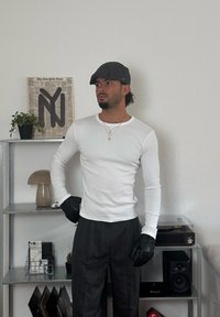 Fehér, hosszú ujjú, testhezálló ing kerek nyakkivágással, fekete kesztyűk és sötétszürke nadrág. A háttérben növény és dísztárgyak láthatók.