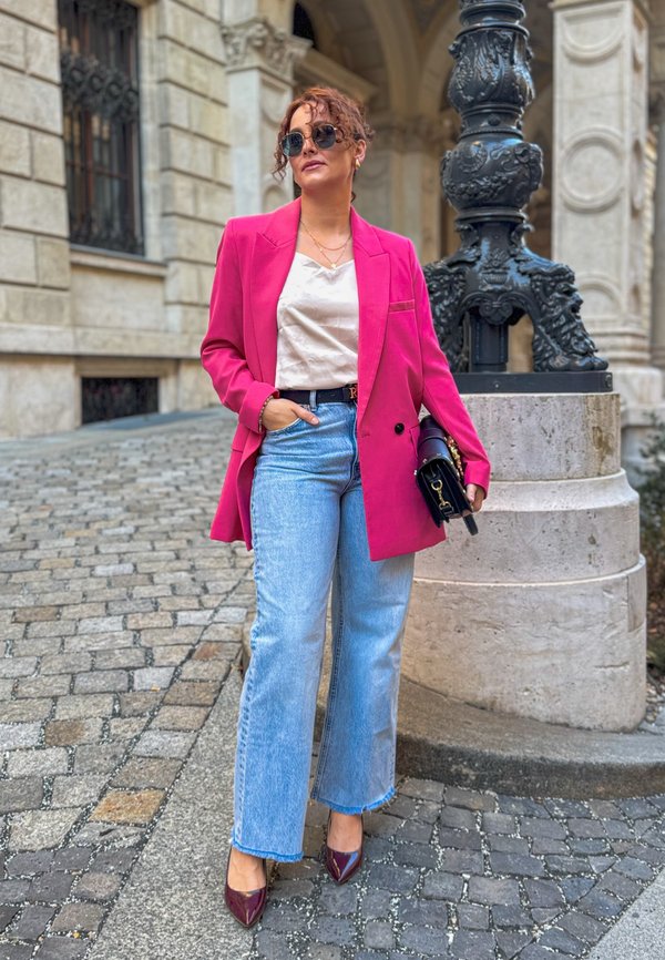 Bright pink jacket over a beige blouse, paired with light blue wide-leg jeans and burgundy high heels. Accessorized with sunglasses and a black bag.