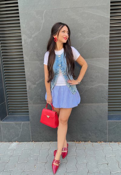 Chaleco de denim sobre una camiseta blanca, falda a rayas azules, bolso rojo y zapatos de punta rojos con tachuelas; de pie frente a una pared gris.