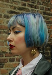 Κοντό καρέ κούρεμα με παστέλ μπλέ και πράσινες αποχρώσεις. Διαθέτει ένα εξέχον χρυσό σκουλαρίκι σε σχήμα καρδιάς, έντονο μπλε eyeliner και κόκκινα χείλη.