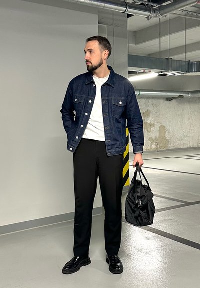 Chaqueta de mezclilla en azul oscuro, camisa blanca, pantalones negros y zapatos negros. Sosteniendo un bolso negro. Fondo neutro con tonos grises.